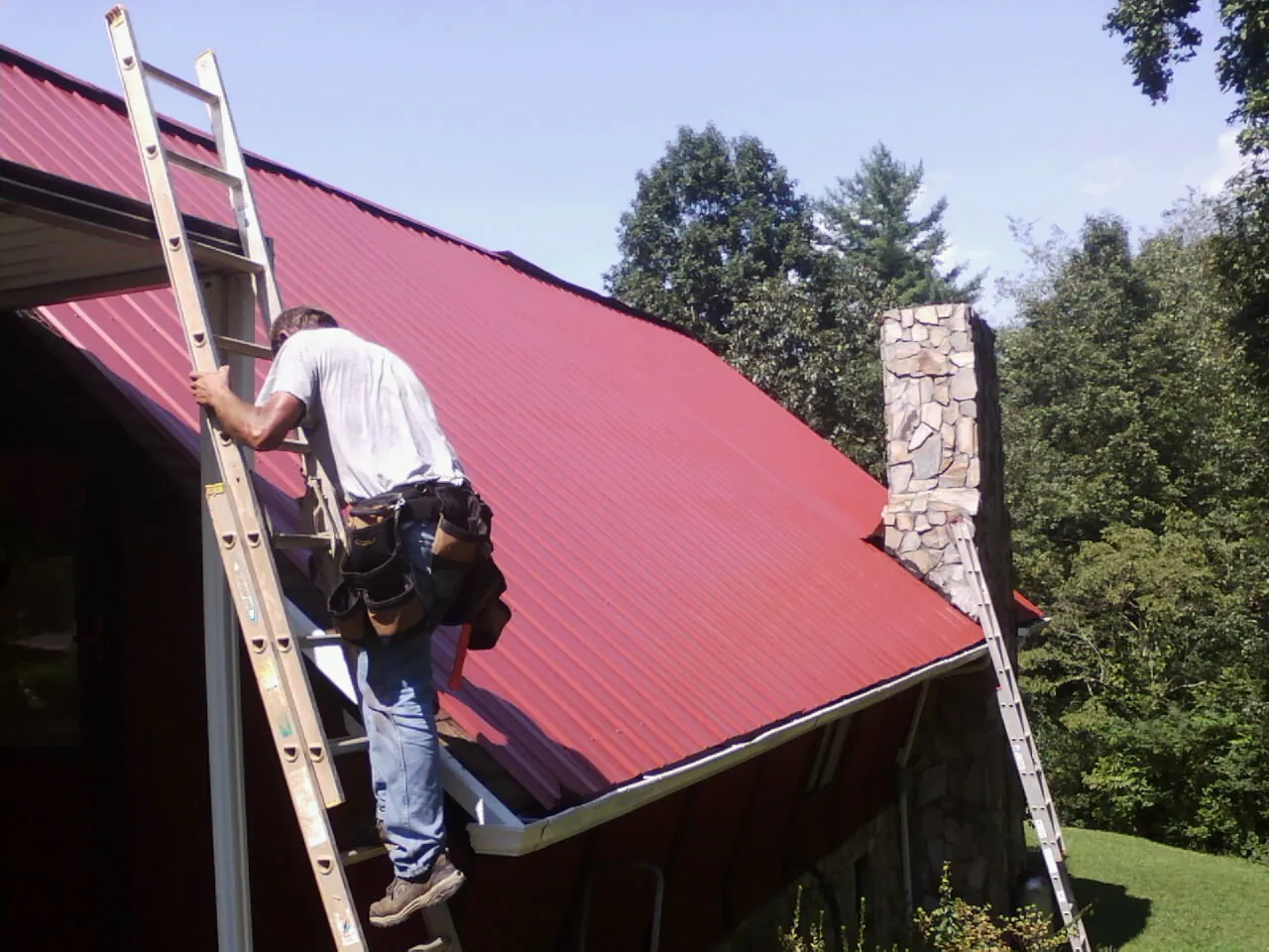 Roofer on a ladder beside a red metal roof and stone chimney — Soffit & Fascia Repair by Roofing Brothers in Lindstrom